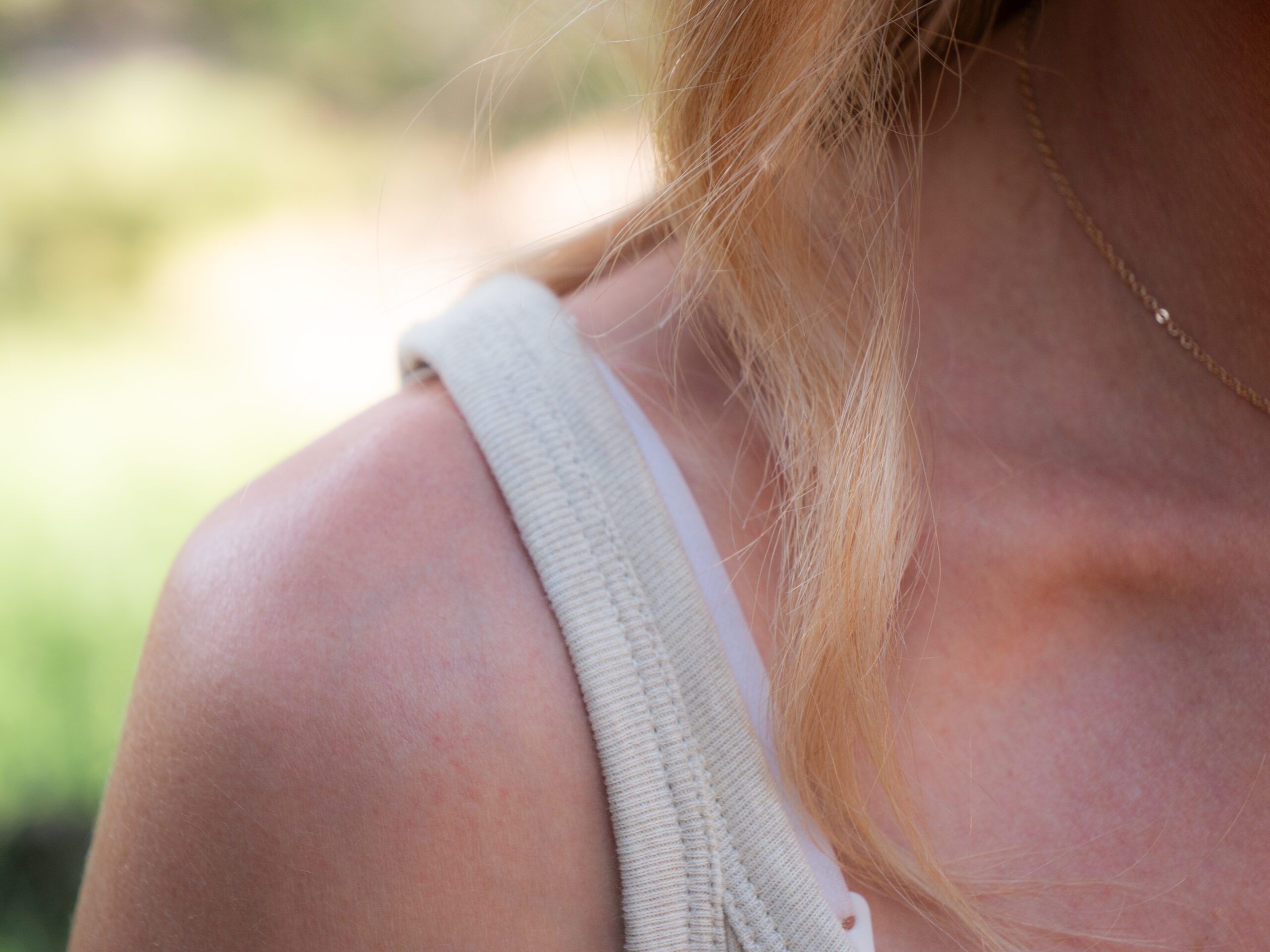 Close-up of a person's shoulder and neck with light skin, wearing a beige tank top and a delicate gold necklace, with blonde hair partially visible.