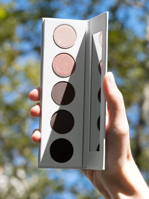 A hand holds an open eyeshadow palette with five neutral shades and a mirror, with trees and sky blurred in the background.