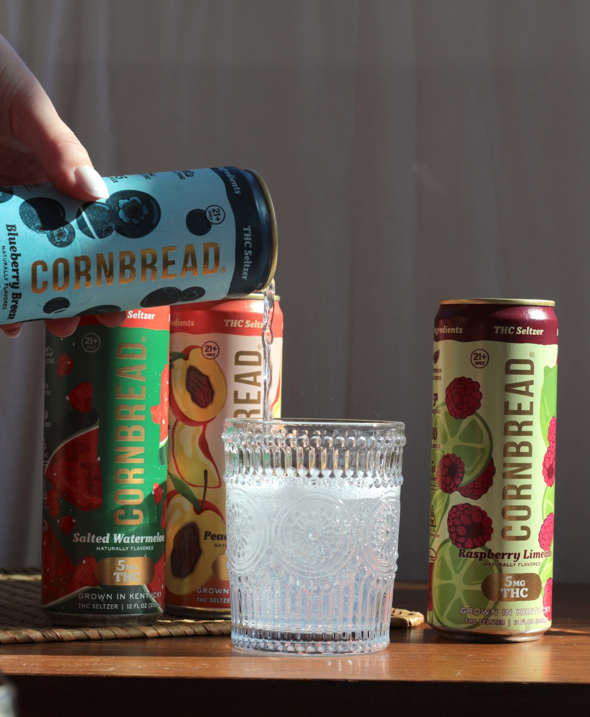 A hand pours a Cornbread THC seltzer into a clear textured glass, with three other Cornbread seltzer cans lined up beside it on a wooden surface.