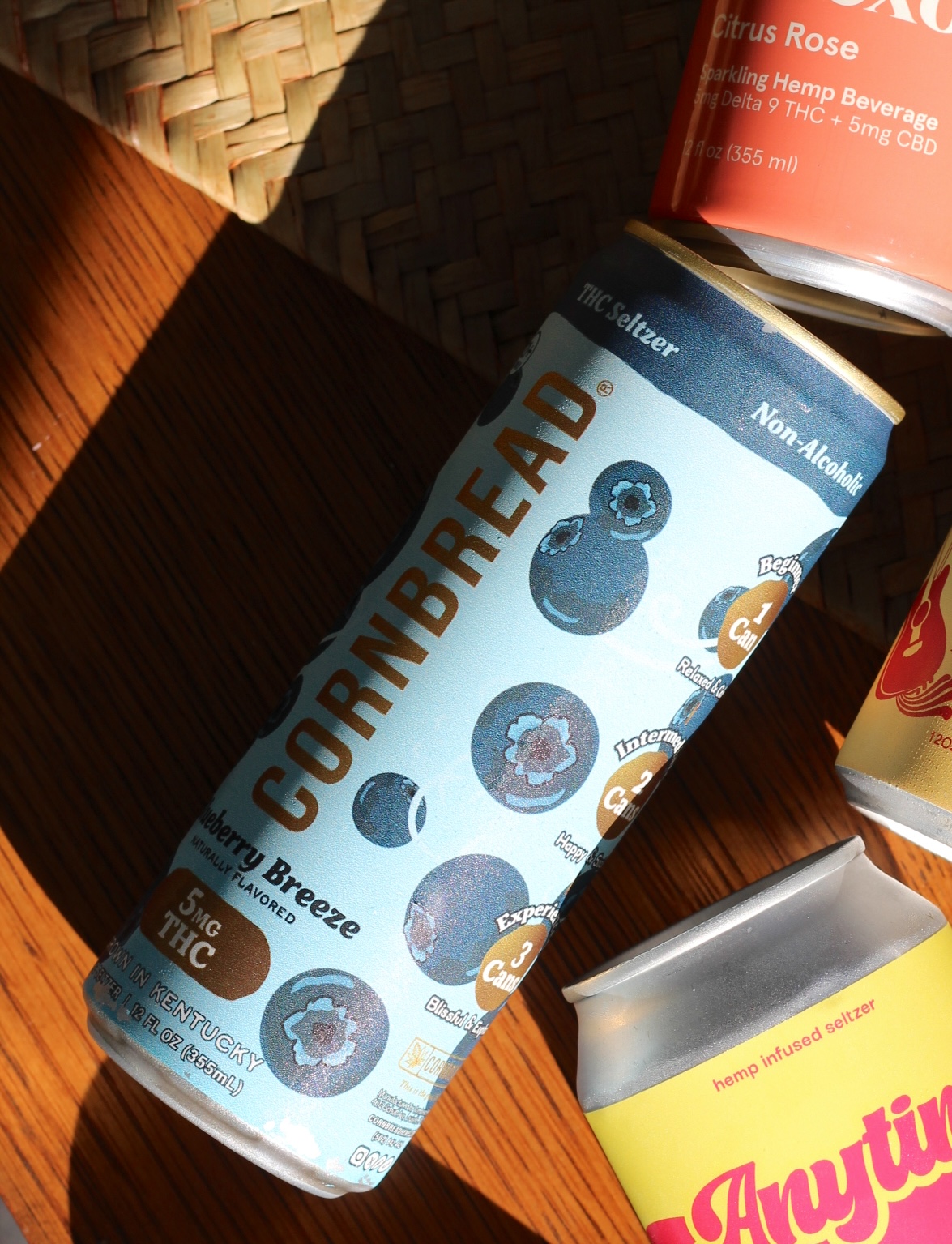 A can of Cornbread Blueberry Breeze THC seltzer lies on a wooden surface, surrounded by other colorful beverage cans in sunlight.