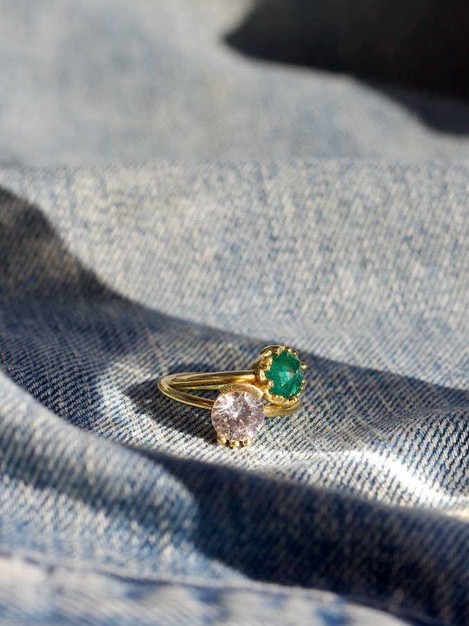 A gold ring with two stones, one green and one clear, is placed on blue denim fabric in natural light.