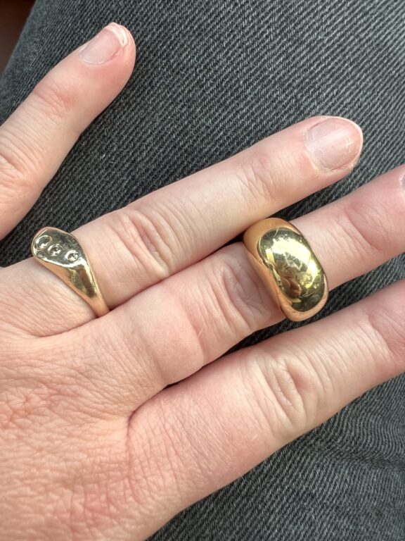 A hand with two gold rings on the middle and ring fingers, shown against blue denim fabric. The nails are natural and unpolished.