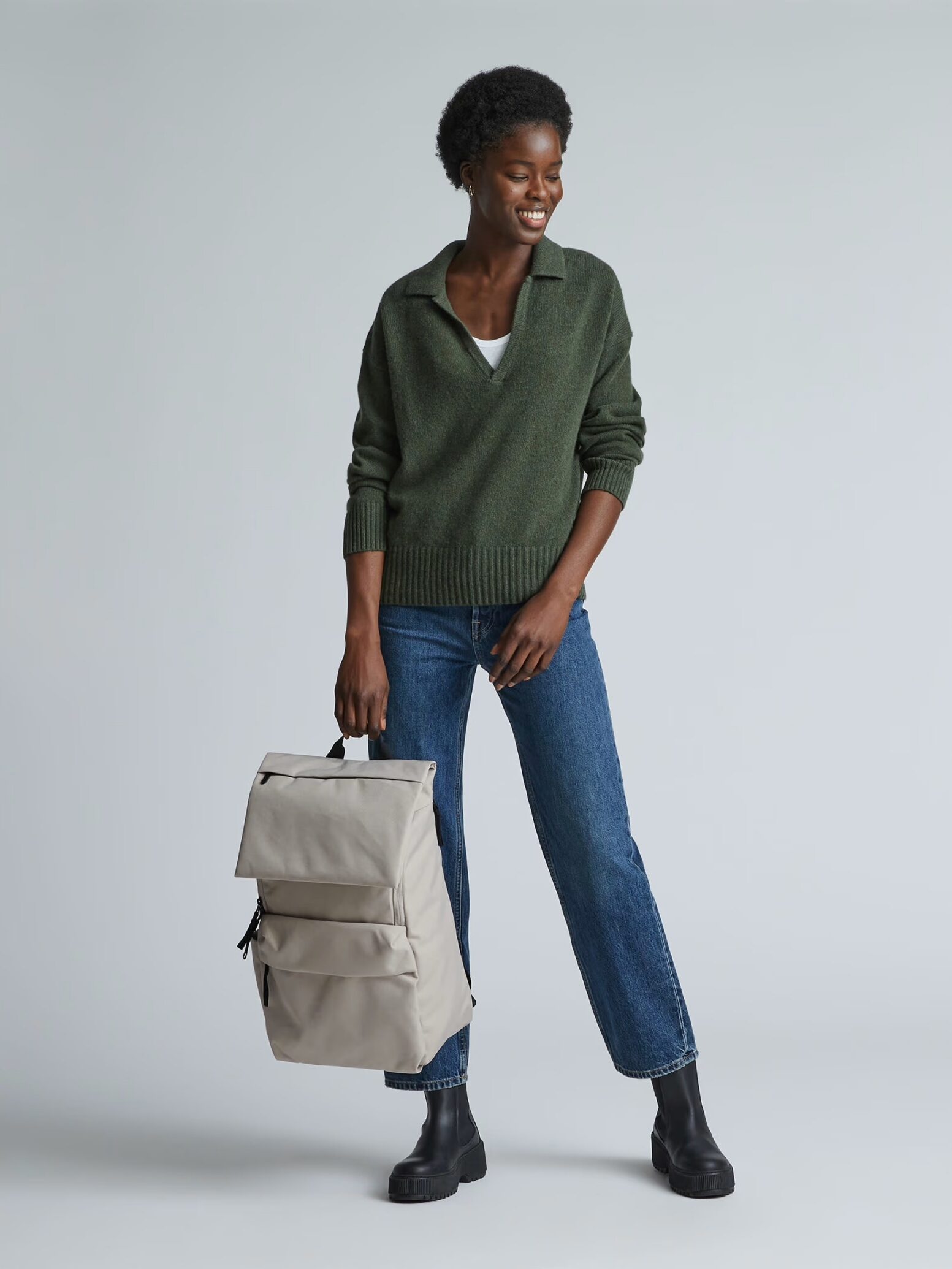 A person wearing a green sweater, blue jeans, and black boots stands holding a beige backpack against a plain background.