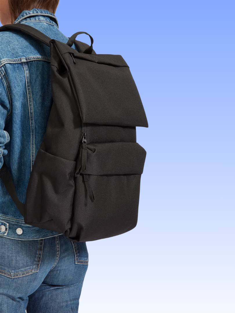 Person wearing a black, rectangular backpack with multiple zippered pockets, dressed in a blue denim jacket and blue jeans, against a gradient blue background.