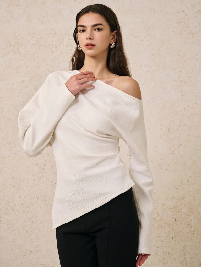 Woman wearing a textured off-shoulder white top with long sleeves and black pants, channeling effortless French girl fashion against a beige speckled background.
