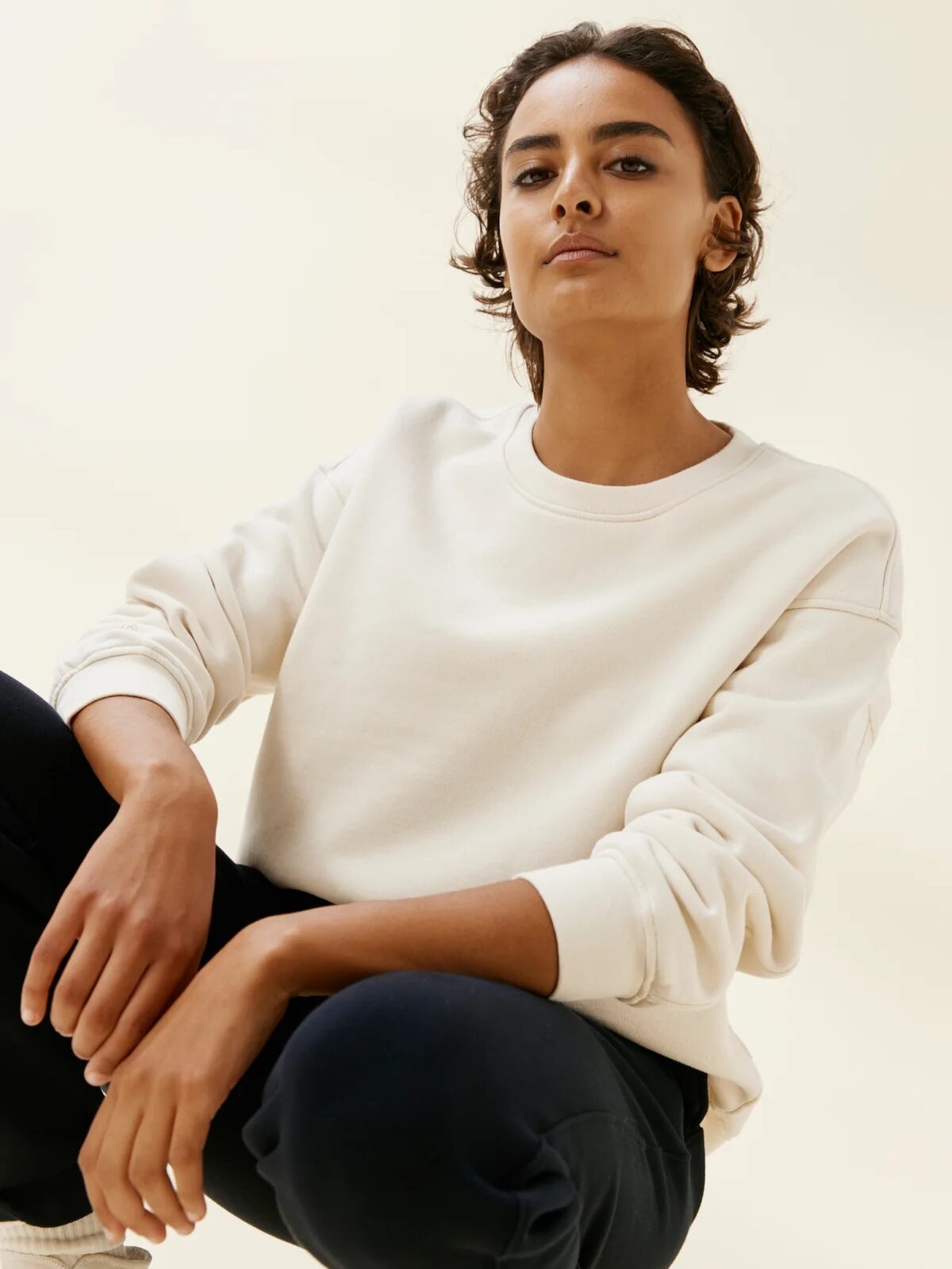 A person channels french girl fashion in a cream sweatshirt and dark pants, sitting with one knee up against a plain light background.