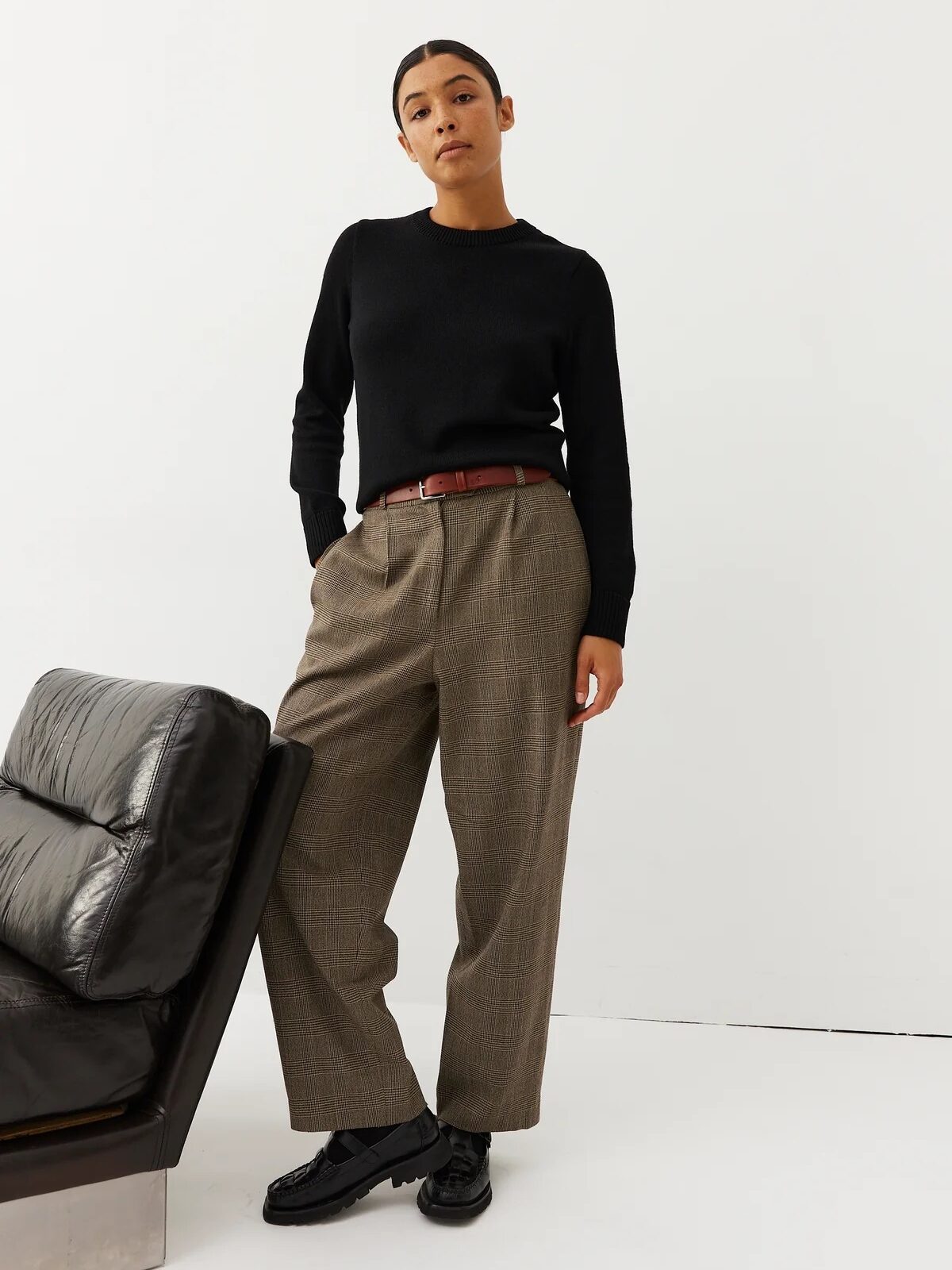 A person embodies French girl fashion indoors, wearing a black sweater, brown wide-leg trousers with a belt, and black shoes, leaning slightly against a black leather chair.