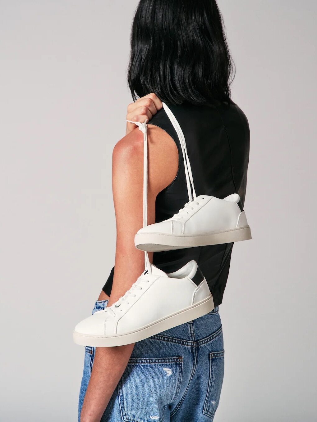 Channeling effortless french girl fashion, a person in a sleeveless black top and blue jeans casually holds a pair of white sneakers by the laces over their shoulder.