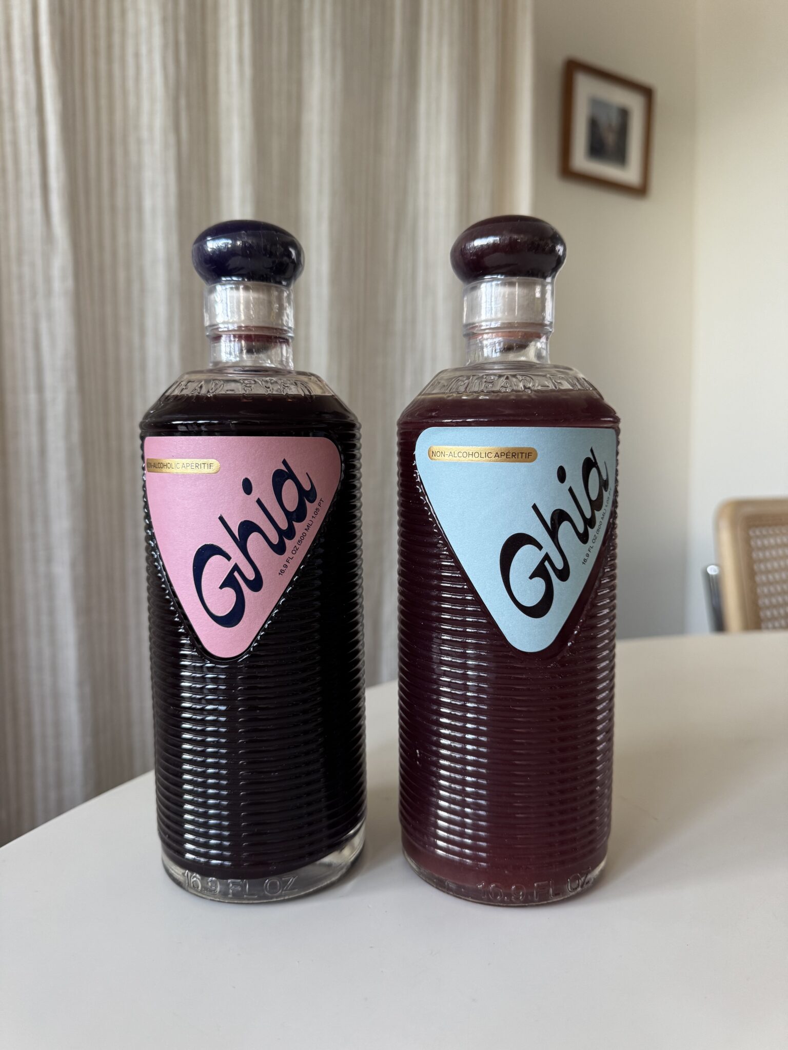 Two bottles of Ghia non-alcoholic aperitif sit side by side on a white table, one with a pink label and one with a blue label, in a room with a curtain and framed photo on the wall.