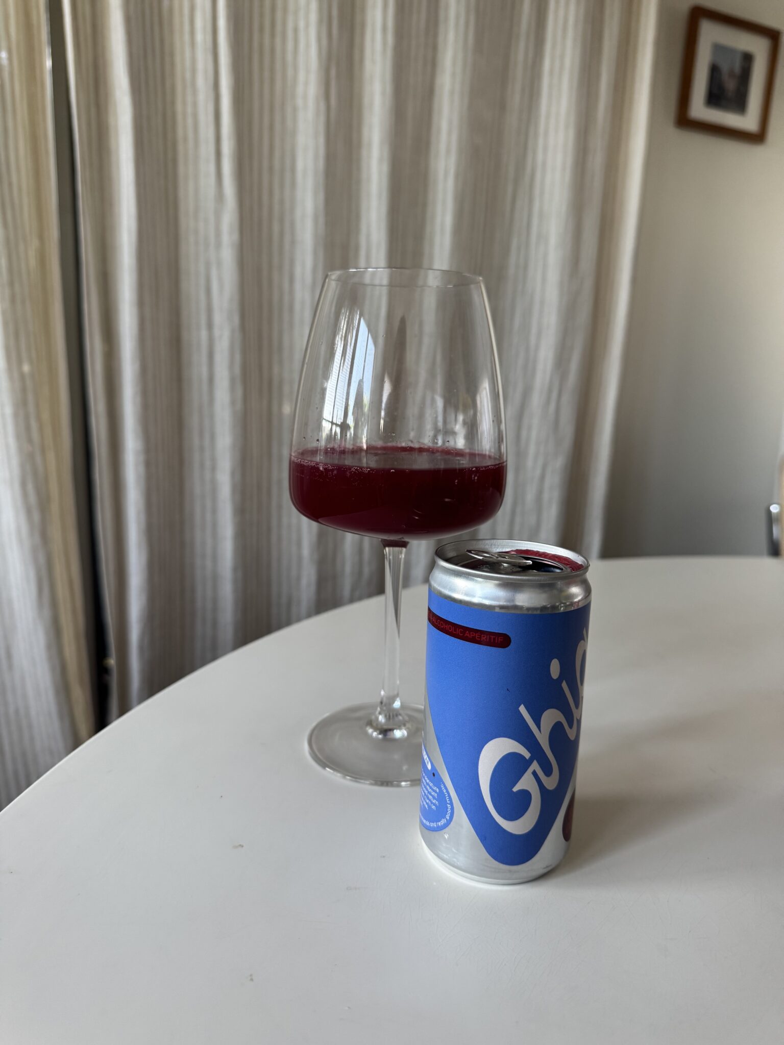 A glass of dark red beverage and a blue can labeled "Ghia" are placed on a white round table in front of cream-colored curtains.