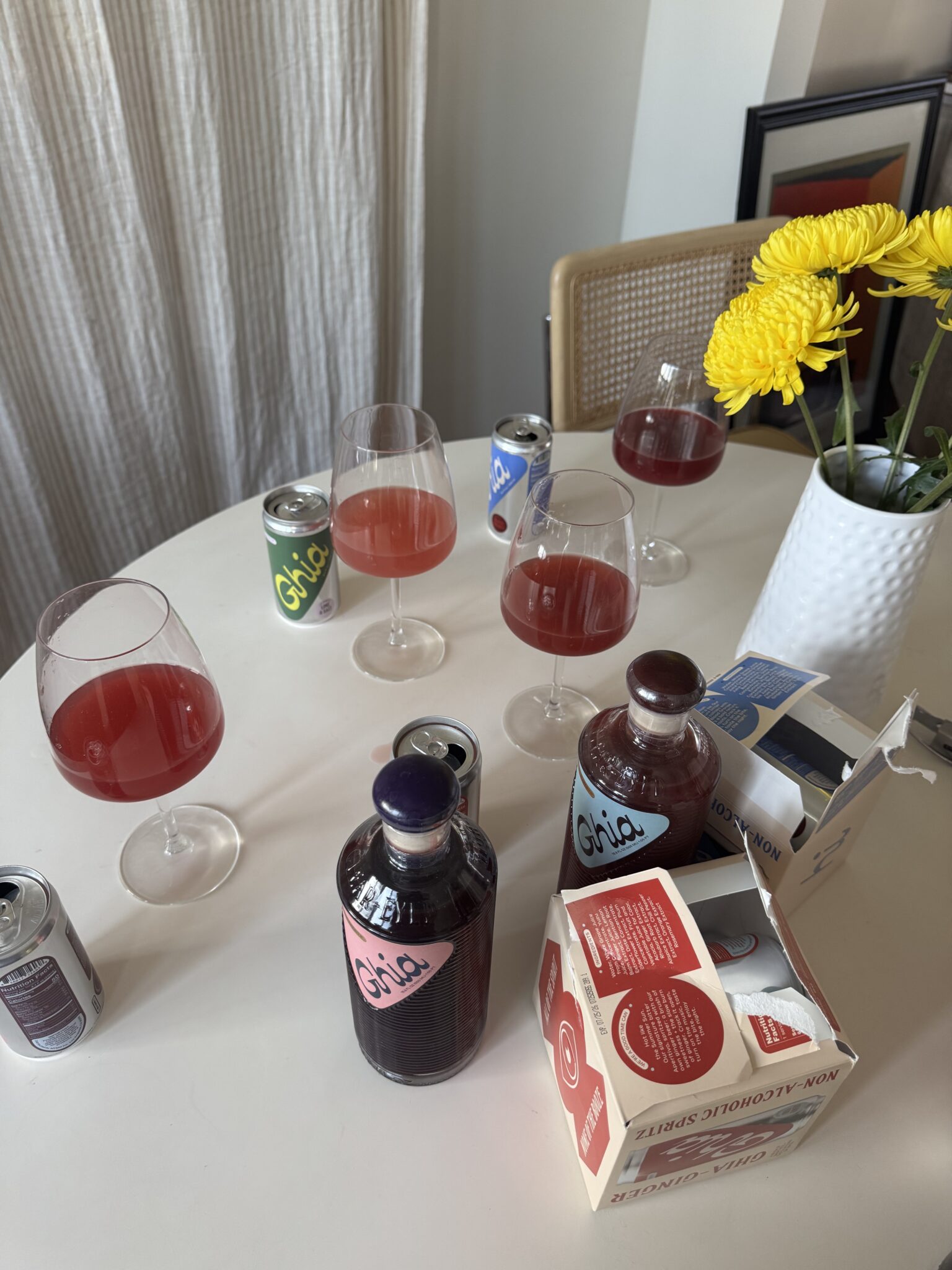 A round table with three glasses of red drink, non-alcoholic spirit bottles, cans, a carton, and a white vase with yellow flowers.