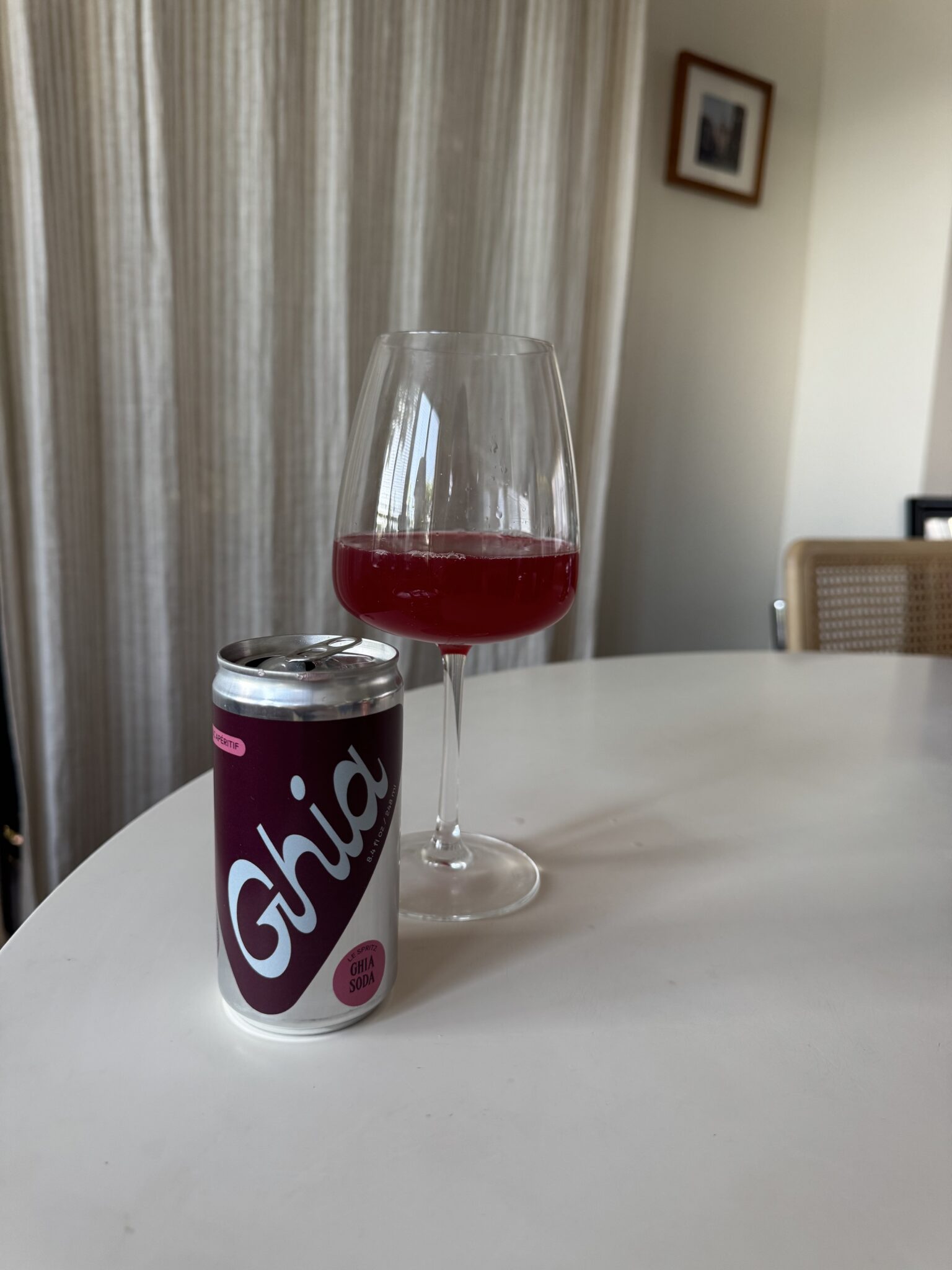 A can of Ghia soda and a wine glass filled with a red beverage sit on a white table in a room with curtains and a framed picture on the wall.