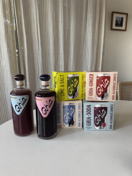 Two glass bottles and four boxes of Ghia non-alcoholic beverages are arranged on a white surface in front of beige curtains.