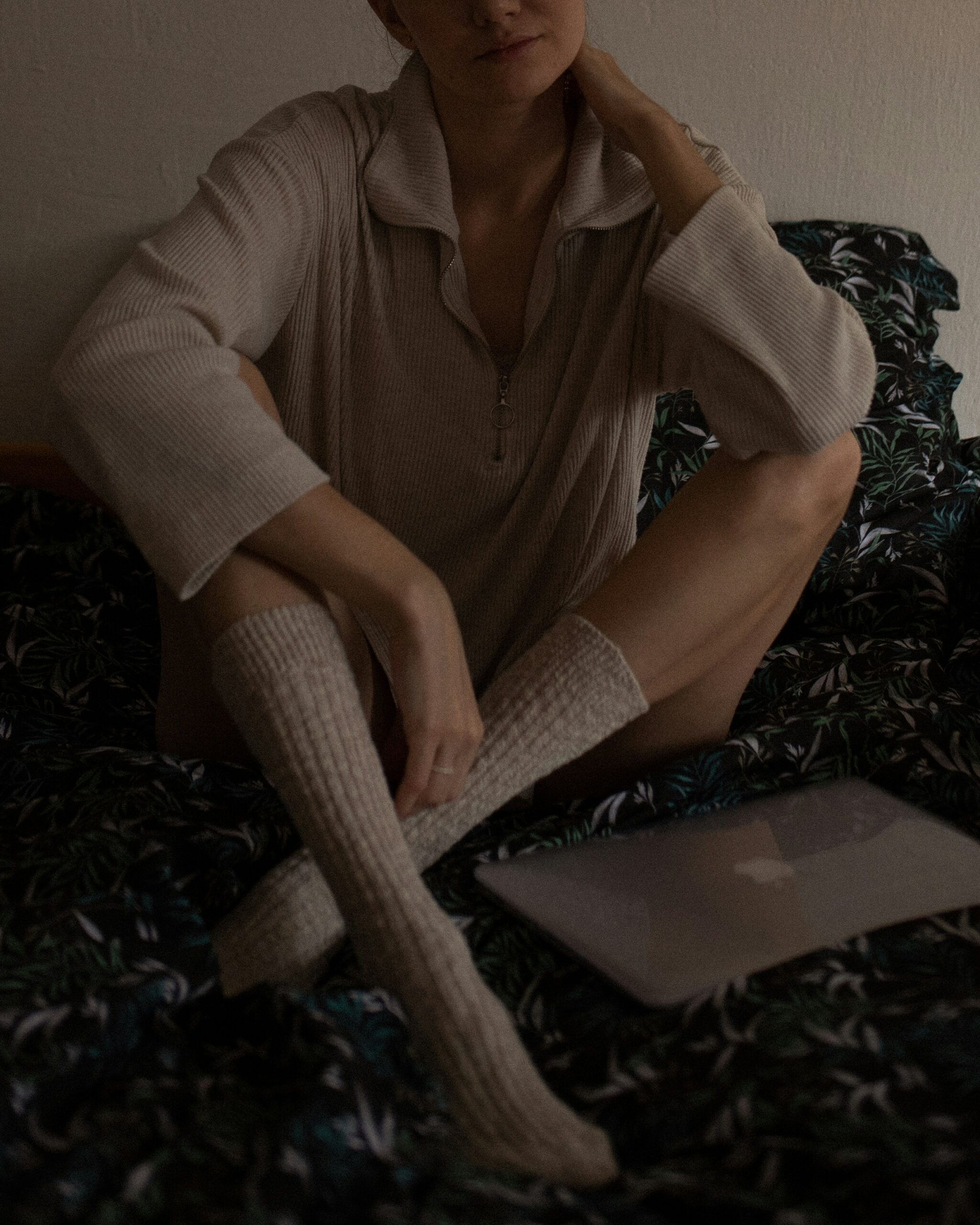 Person sitting cross-legged on a bed wearing a long-sleeve top and knee-high socks, with a closed laptop resting on the bed beside them.