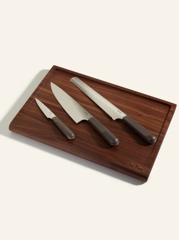 A wooden cutting board with three knives: a paring knife, a chef’s knife, and a serrated bread knife, all with dark handles, displayed on a light background.