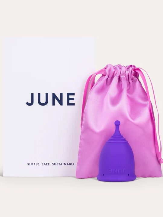 A purple menstrual cup beside a pink drawstring pouch and a white card with "JUNE" written on it.