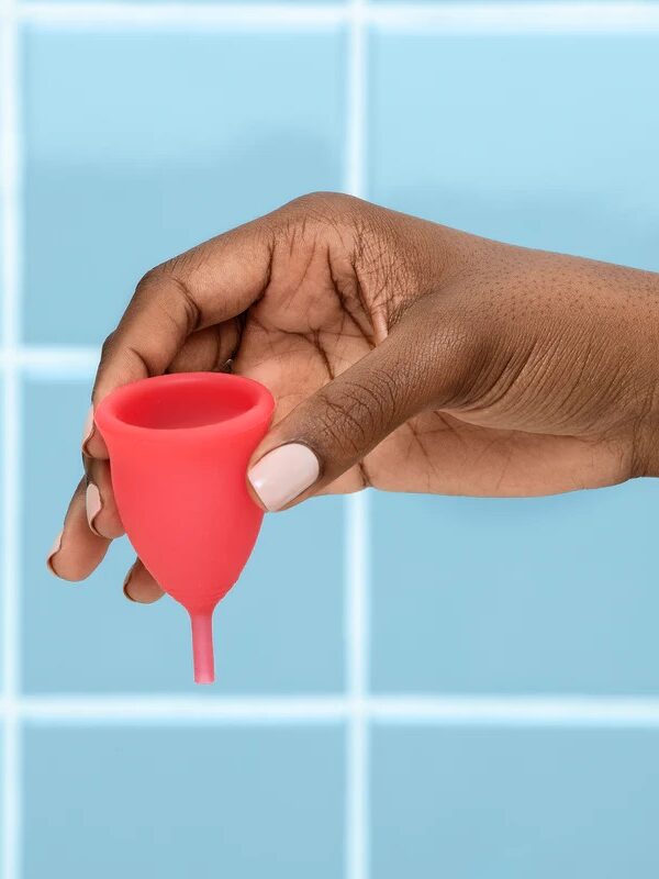 A hand holding a red menstrual cup in front of a blue tiled background.