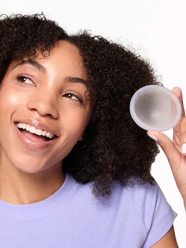 A person with curly hair, wearing a lavender shirt, smiles and holds up a clear circular object in their right hand against a plain background.