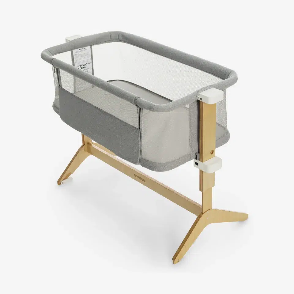 A modern bassinet with a grey fabric and mesh design, supported by a light wooden frame with angled legs, shown on a white background.