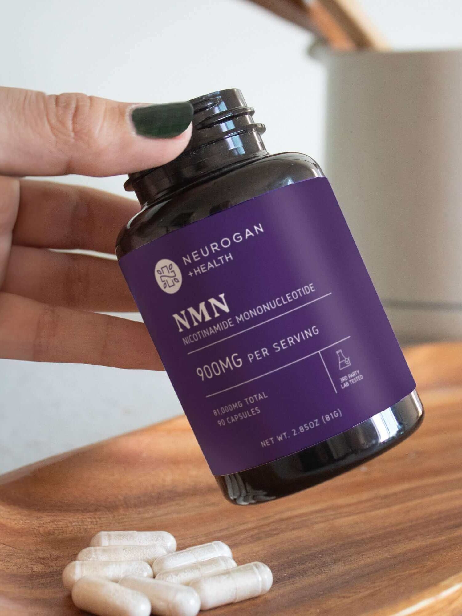 A hand holds a bottle of Neurogan NMN 900mg, one of the best NMN supplements, with capsules displayed on a wooden tray in the foreground.