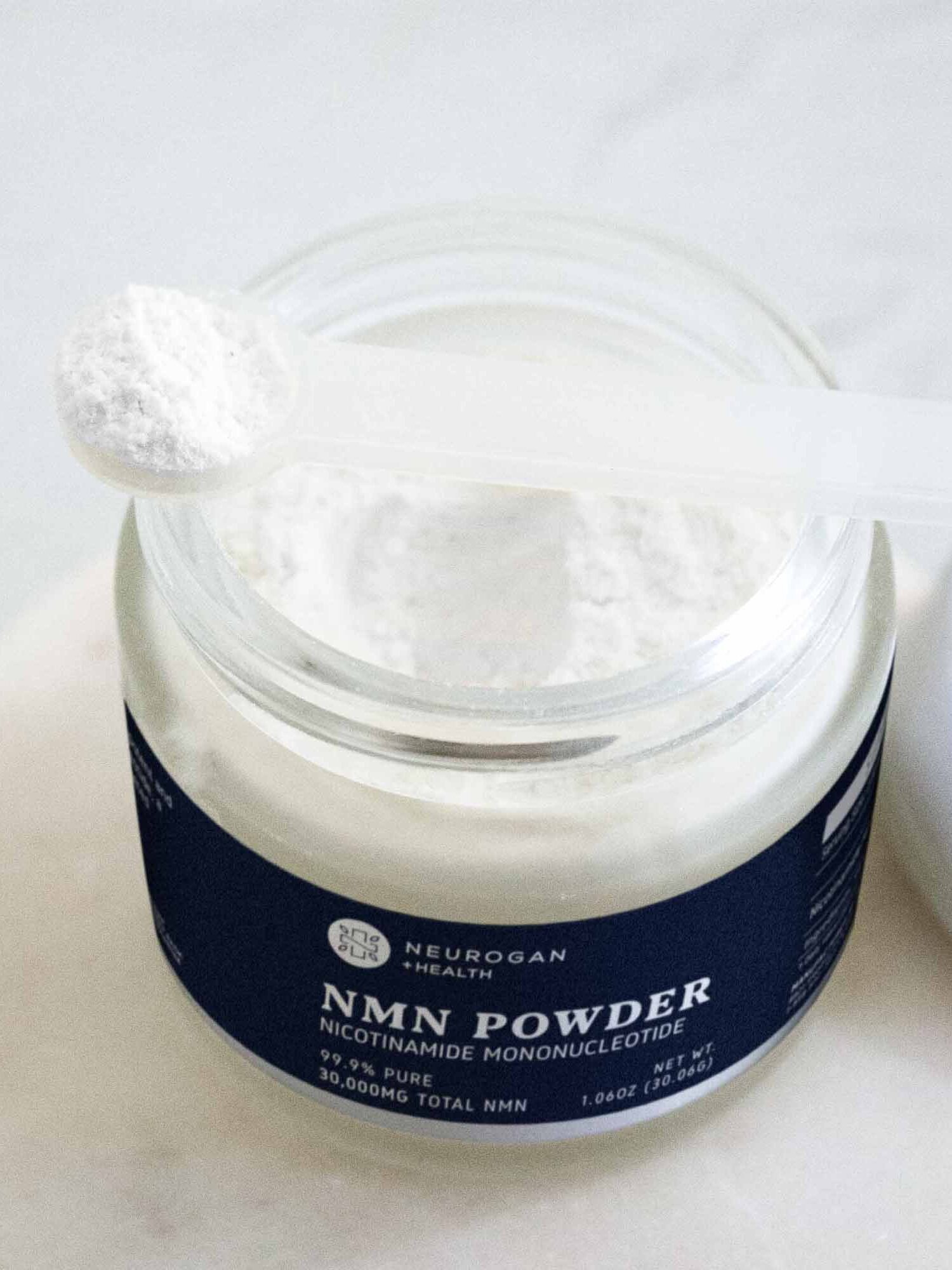 A plastic scoop with white powder rests above an open jar labeled “NMN Powder Nicotinamide Mononucleotide,” representing a top choice for those seeking the best NMN supplement, displayed on a clean white surface.