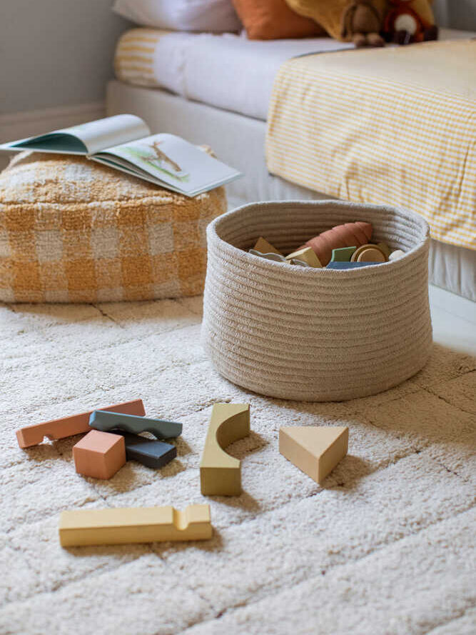 A soft rug with scattered wooden toy blocks, a woven basket, a checkered pouf with an open book, and a neatly made bed with plush toys and a yellow blanket in the background.