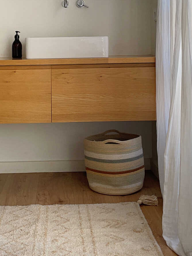 A modern bathroom vanity with a white rectangular sink, wall-mounted faucet, soap dispenser, and a striped woven basket underneath. A white curtain and rug are also visible.
