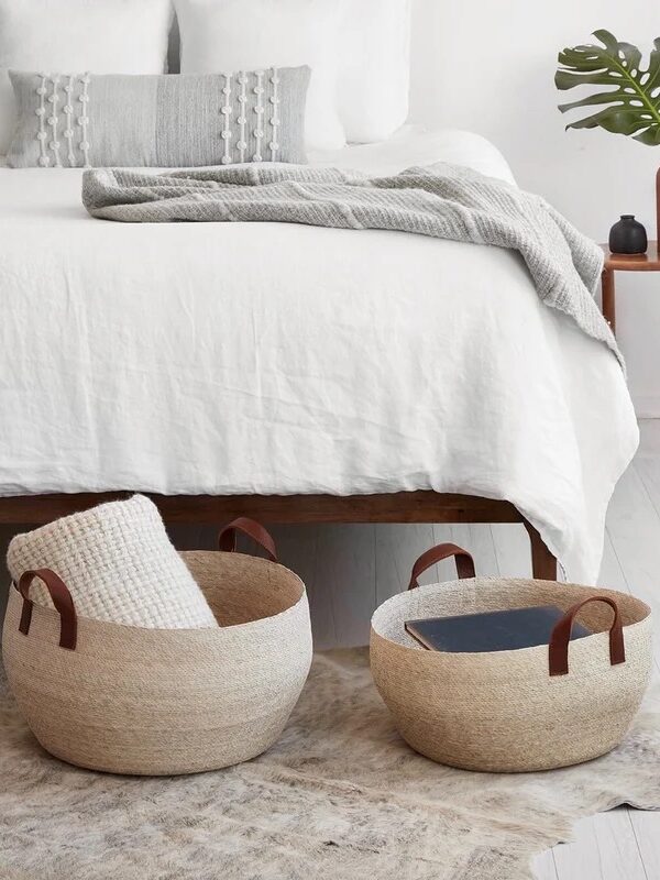 A minimalist bedroom with a white bedspread, two woven baskets on the floor—one holding a blanket, the other a book—and a wooden side table with a vase and green leaf.