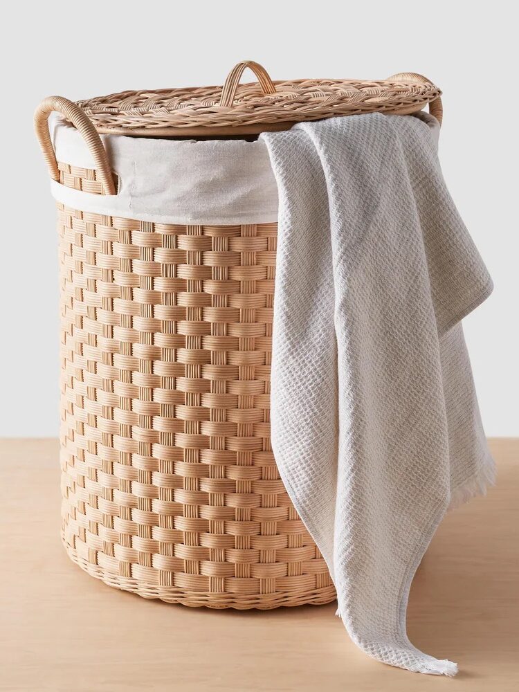 A round woven laundry basket with a lid and side handles sits on a wooden surface, with a light gray towel draped over the side.