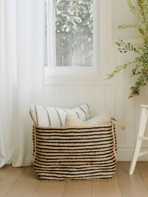 A striped woven basket with cushions sits beside a white chair holding a vase of green branches in a bright, airy room with a window and light wood floor.