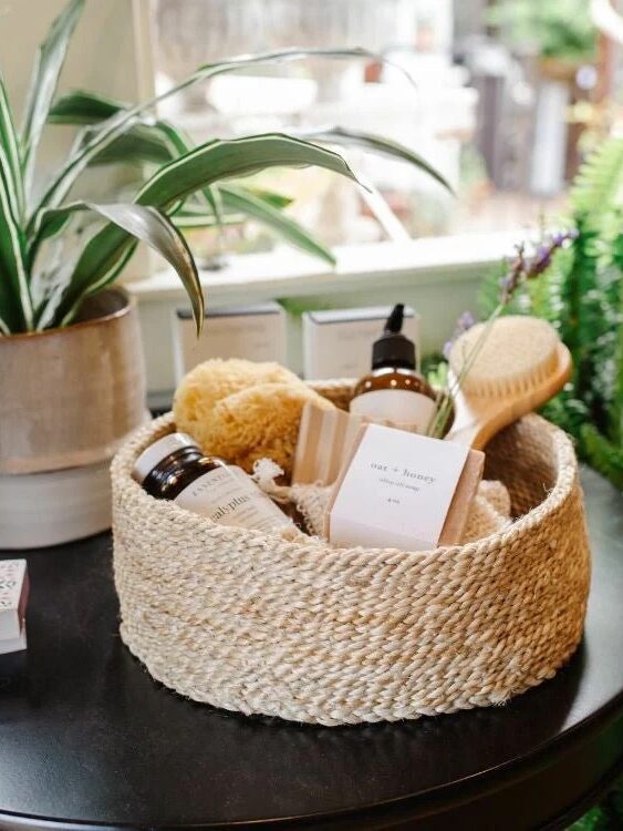 A woven basket on a table holds skincare products, a sponge, a wooden brush, and a bar of soap. A potted plant sits beside it and ferns are in the background.