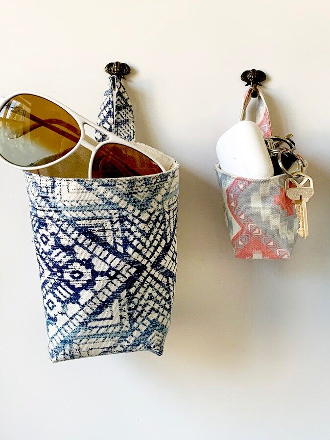 Three patterned fabric wall pouches hang on hooks, holding items such as sunglasses, keys, lip balm, and sunscreen against a white background.