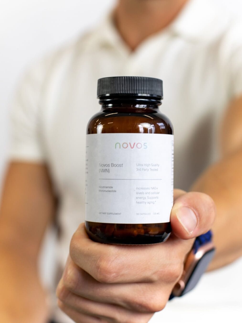 A person wearing a white shirt holds a brown bottle labeled "Novos Boost" dietary supplement, with the label text in focus.