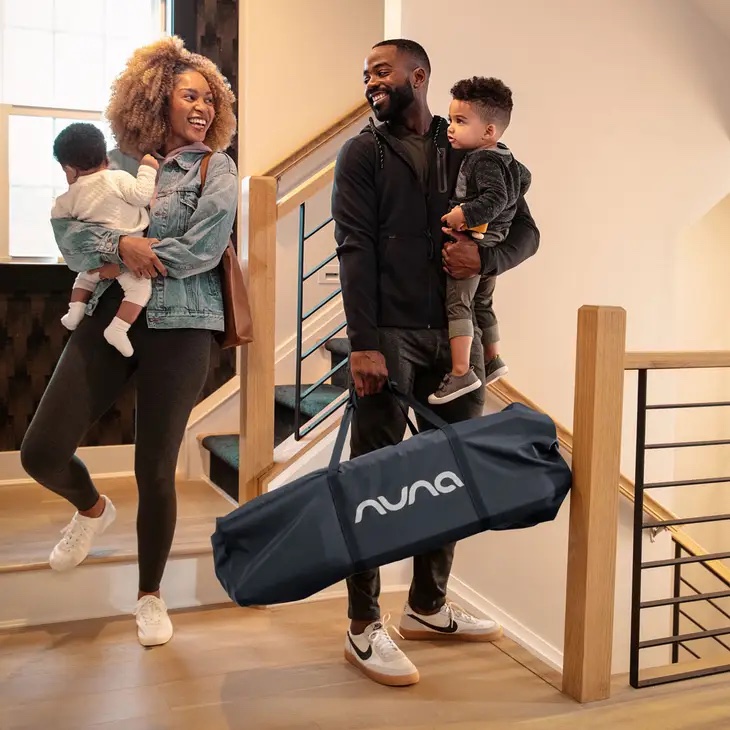 A woman holding a baby and a man holding a toddler stand on stairs; the man carries a large black Nuna bag.