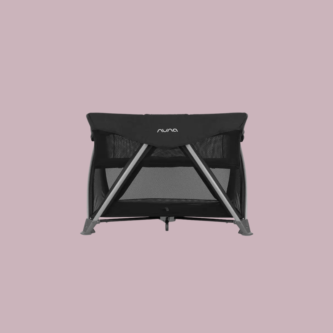 A black Nuna travel crib with mesh sides and a metal frame, shown against a plain light mauve background.