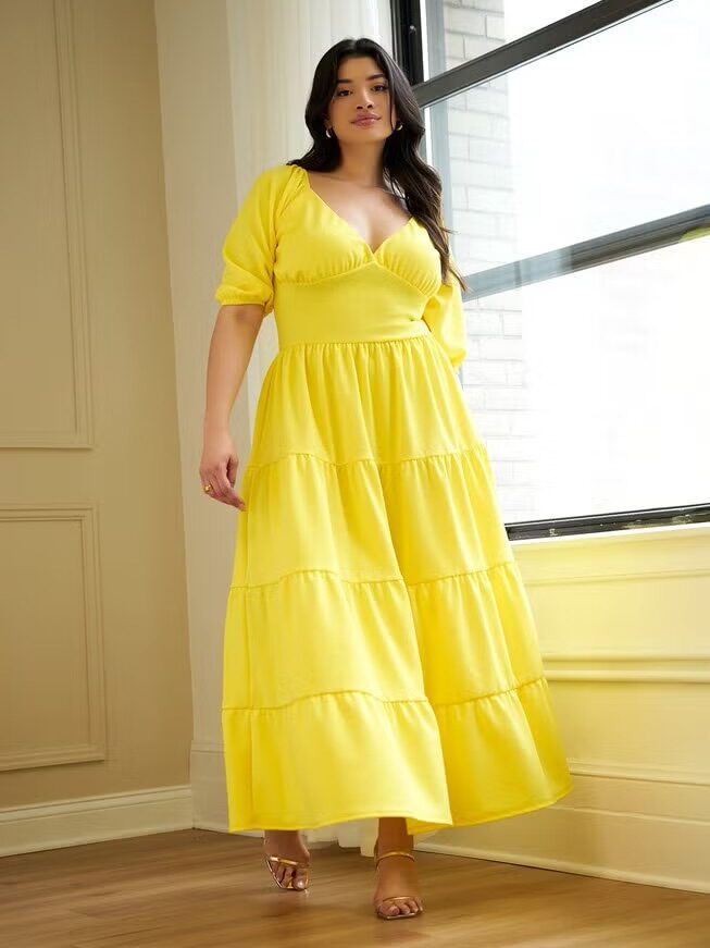 A woman stands indoors wearing a bright yellow, tiered maxi dress with puff sleeves and open-toe heels, beside a large window.