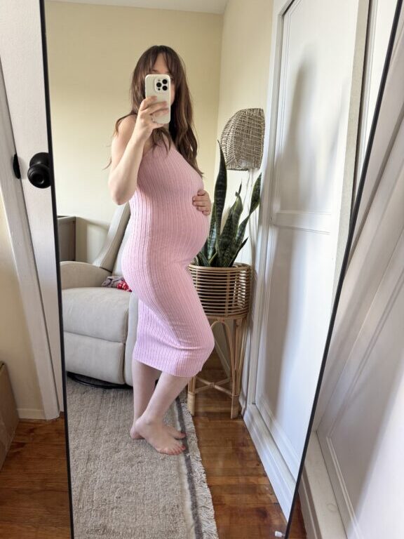A person in a pink ribbed dress takes a mirror selfie, showing a pregnant belly, standing on a rug in a room with a chair and a large potted plant.