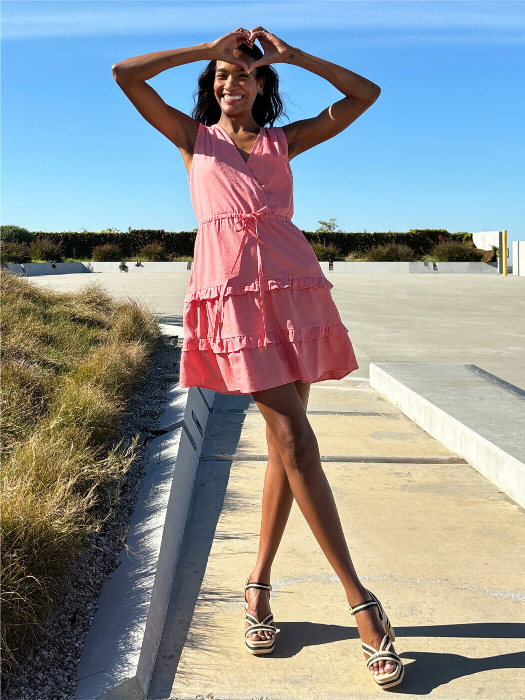 A person in a pink sleeveless dress and sandals stands outdoors on a sunny day, forming a heart shape with their hands above their head.