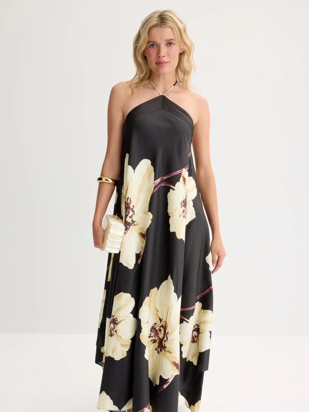 A woman stands against a plain background wearing a black halter-neck maxi dress with large yellow floral prints, gold sandals, a gold bracelet, and holding a small gold clutch.