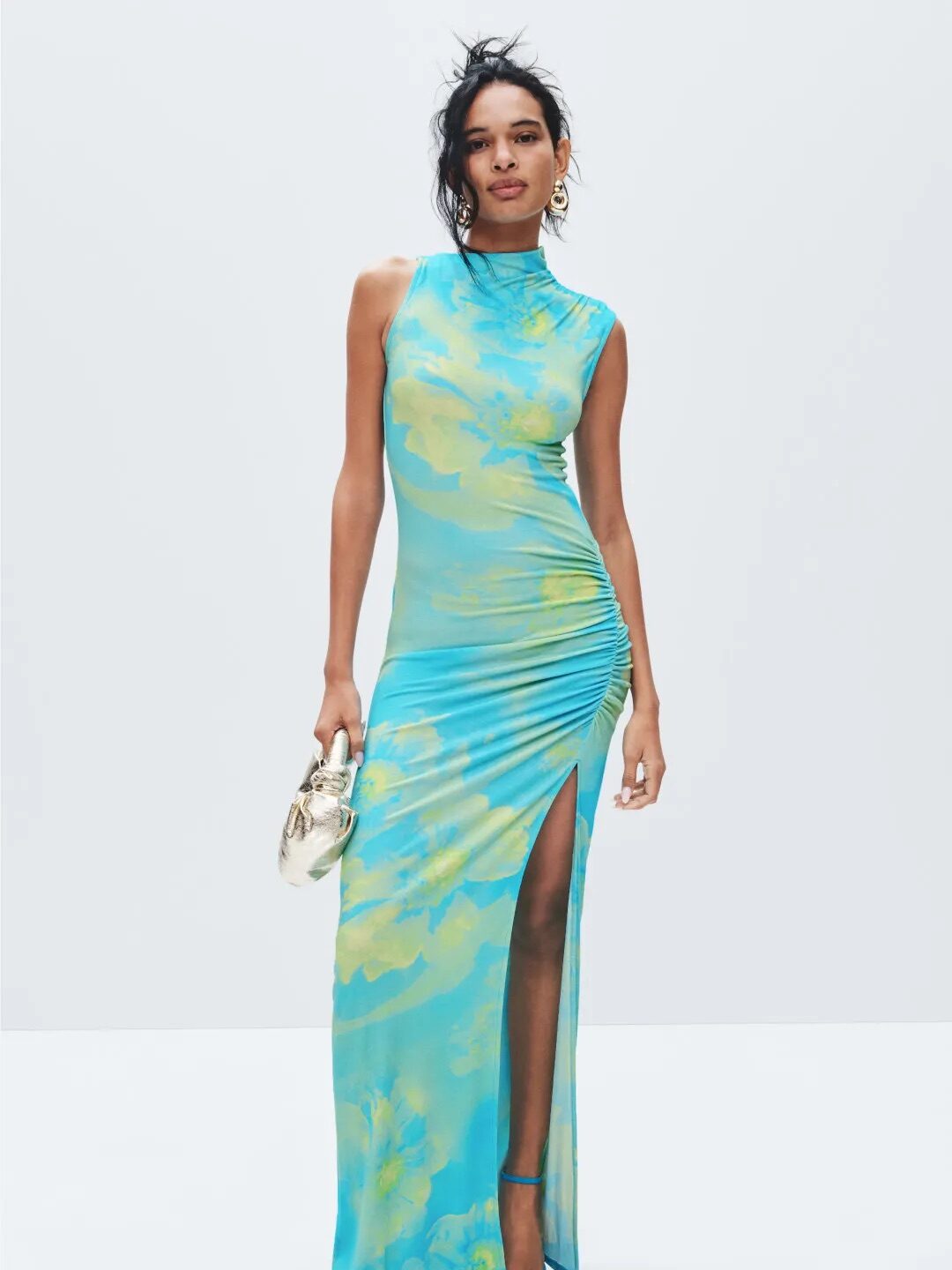 A woman wearing a sleeveless, high-neck, blue and yellow patterned maxi dress with a thigh-high slit, holding a small metallic handbag and wearing blue heeled sandals.