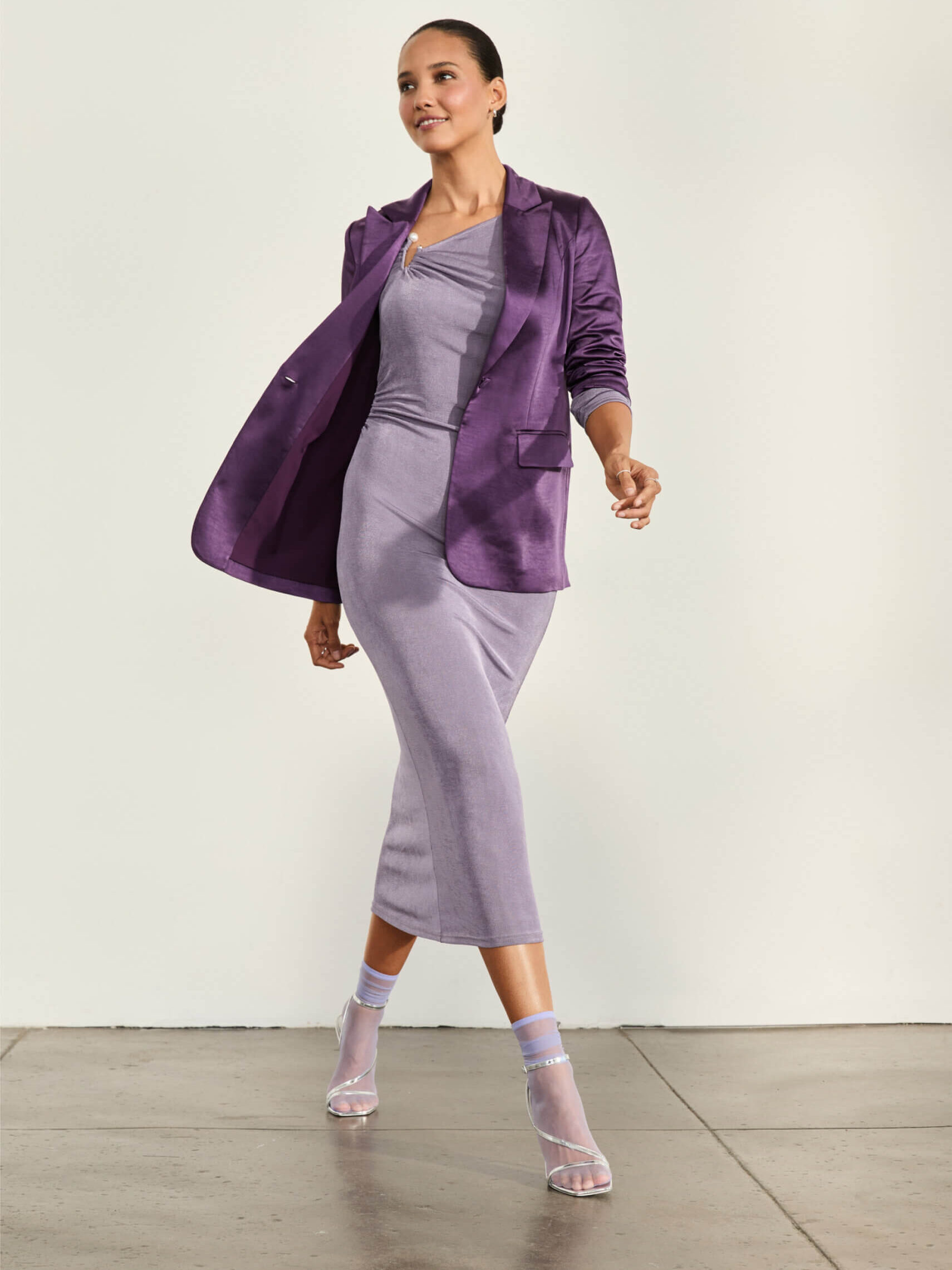 A woman wearing a fitted lavender dress, matching purple blazer, sheer ankle socks, and heels walks indoors on a gray floor against a light background.
