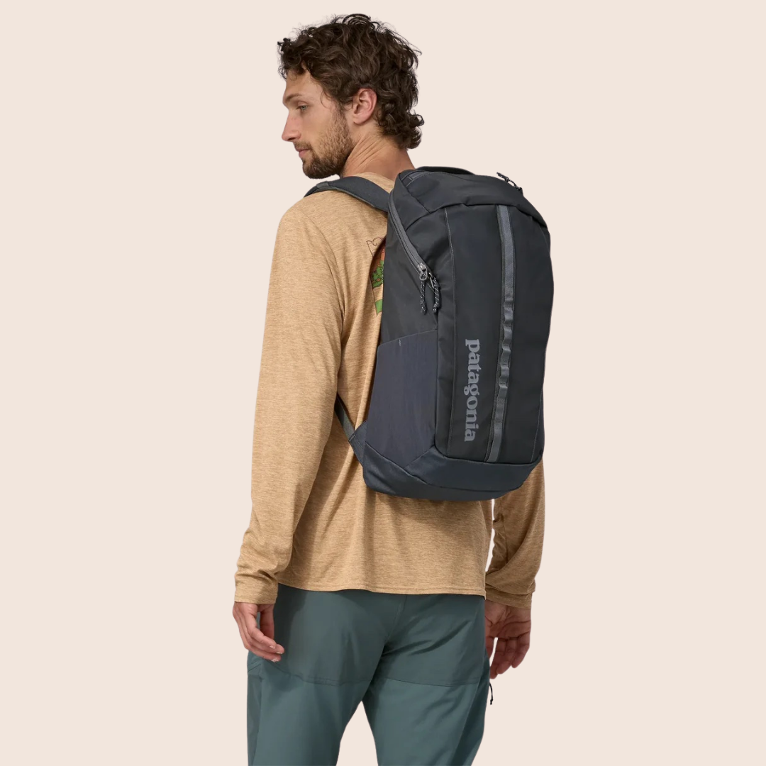 A man wearing a tan long-sleeve shirt and green pants carries a dark gray Patagonia backpack over one shoulder, standing against a plain light background.