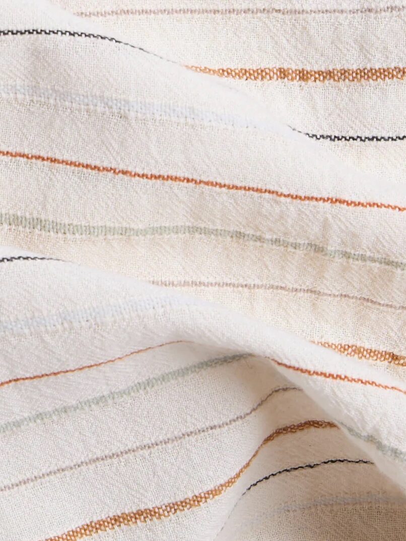 A close-up of a cream-colored fabric with textured, thin horizontal stripes in orange, black, green, and beige. The fabric is slightly wrinkled.