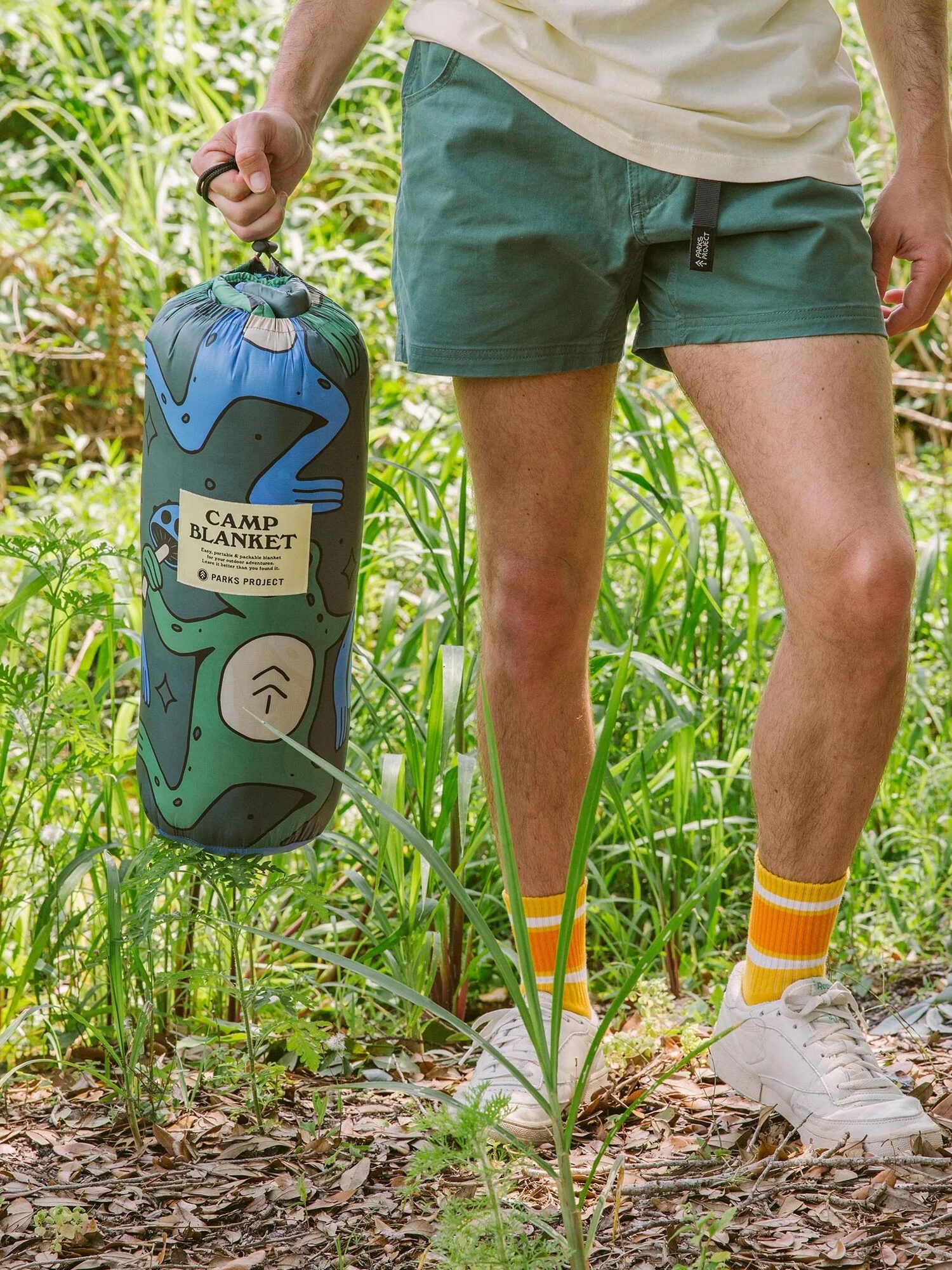 A person wearing teal shorts and yellow socks holds a rolled-up camp blanket in a patterned bag while standing outdoors in a grassy area.