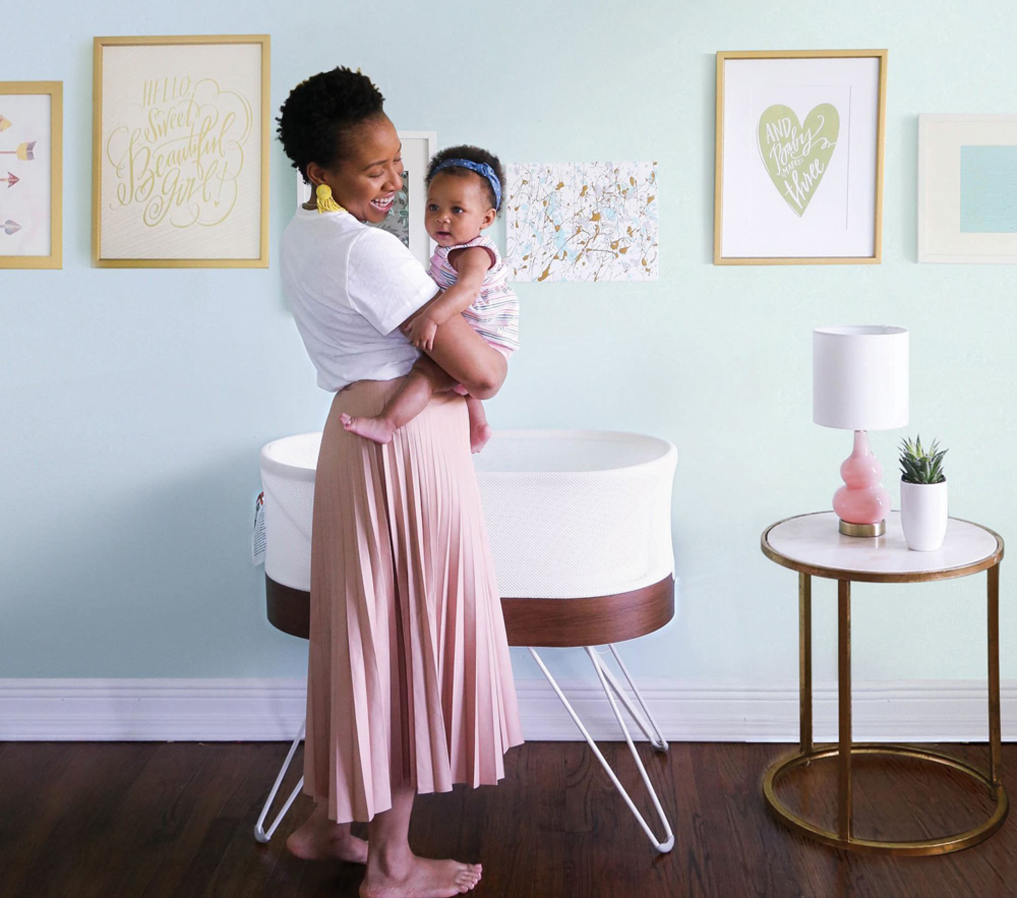 A woman stands in a nursery holding a baby. There is a white bassinet, framed art on the wall, and a small round table with a lamp and decoration.