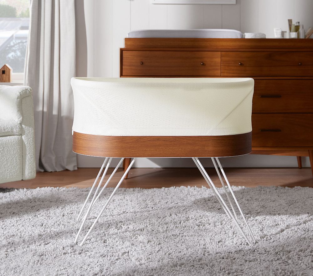 A modern wooden and white bassinet with metal legs sits on a light gray rug in a nursery with a dresser and changing table in the background.