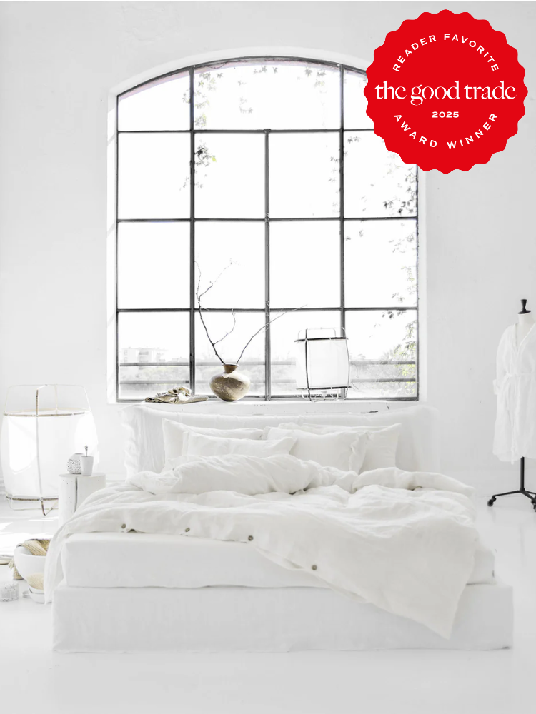 A minimalist bedroom with white bedding, decor, and walls, featuring a large arched window. A red award badge from The Good Trade is overlaid in the top right corner.