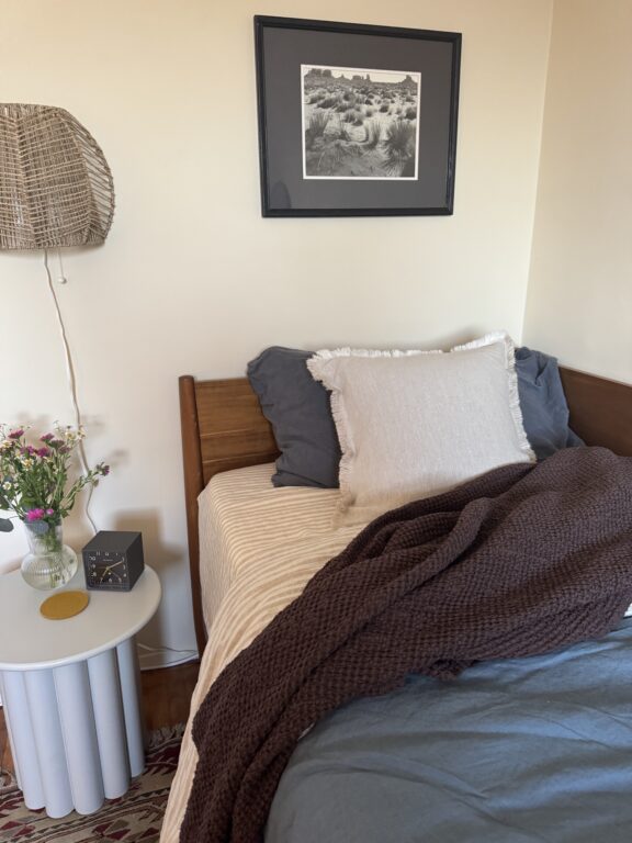A neatly made bed with layered pillows and a brown blanket, next to a round white side table holding flowers, a clock, and a coaster, beneath a framed black-and-white photo.