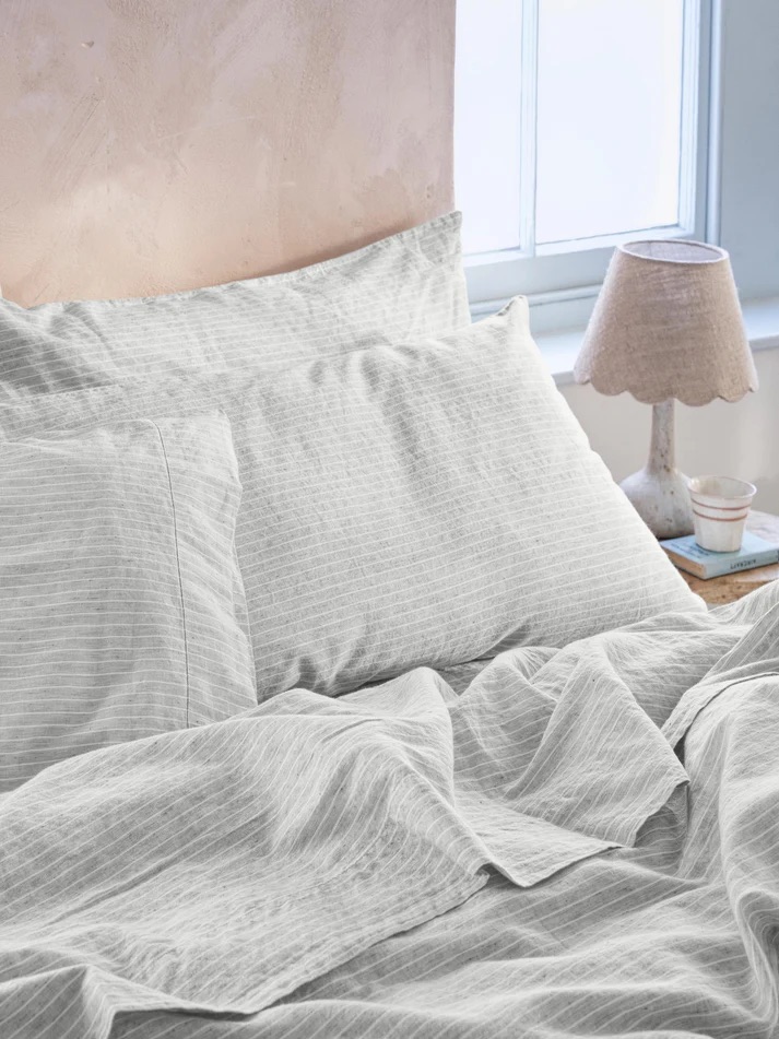 A neatly made bed with light gray striped bedding next to a window and a nightstand with a lamp and a cup.