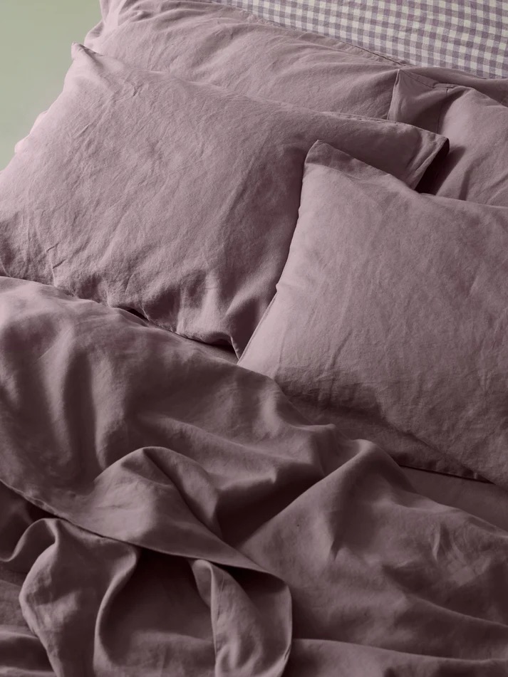 Unmade bed with mauve-colored linen sheets and pillows; a checkered headboard or pillow is partially visible at the top.